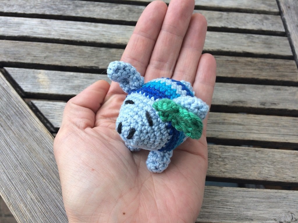 Kleine blau-grüne Amigurumi‑Schildkröte in einer Hand vor Holzlatten