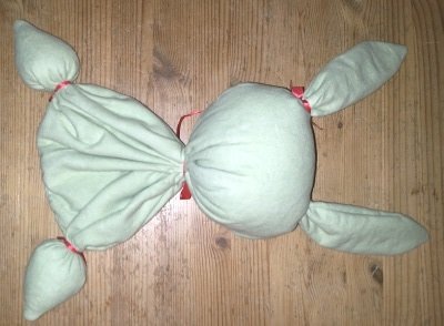 Hellgrünes Babyspielzeug aus Stoff mit roten Bindungen auf einem Holztisch.