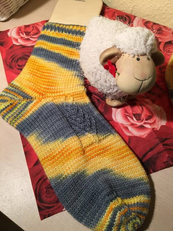 Gestrickte Damensocke in Gelb und Grau auf Sockenbrett neben Schaf-Dekoration.