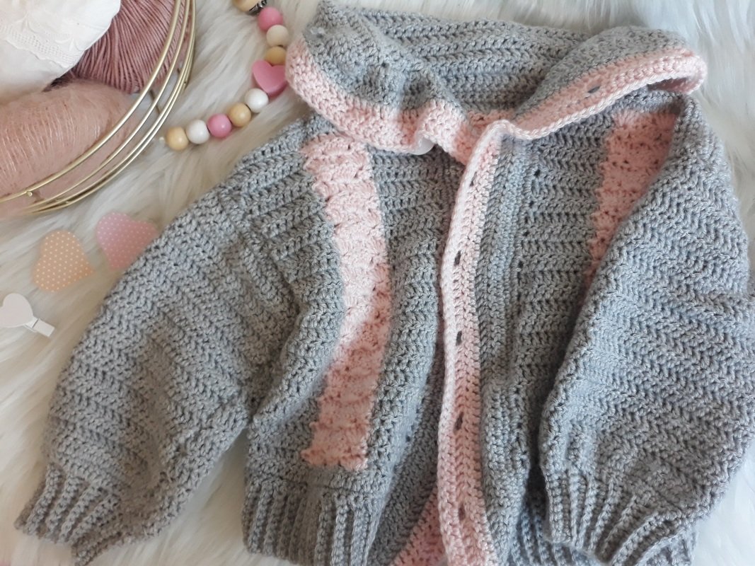 Gehäkelte graue Babyjacke mit rosa Blende und Kapuze, liegend