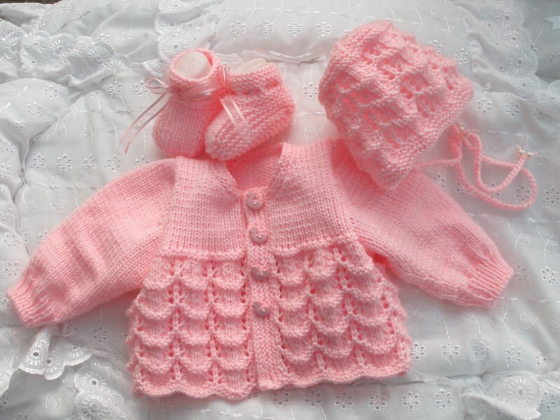 Rosa gestricktes Babyset mit Jäckchen, Mütze und Schühchen mit Muschelmuster auf weißem Stoff