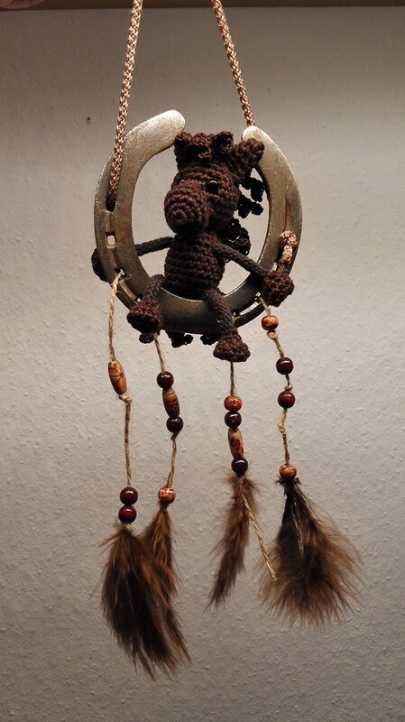 Braunes gehäkeltes Amigurumi-Pferd sitzt in einem Hufeisen, darunter hängen Perlen und braune Federn.