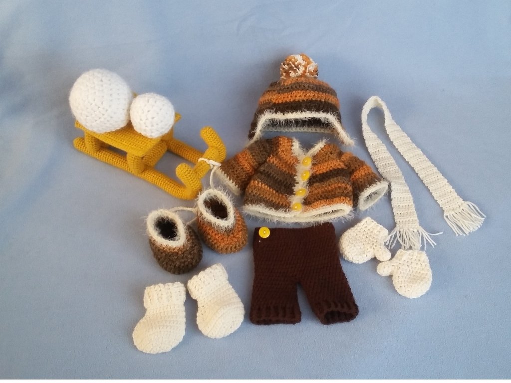 Gehäkeltes Puppen-Winterset: gestreifte Jacke, Mütze, Hose, Schal, Fäustlinge, Stiefel, Socken und gelber Schlitten mit Bällen.