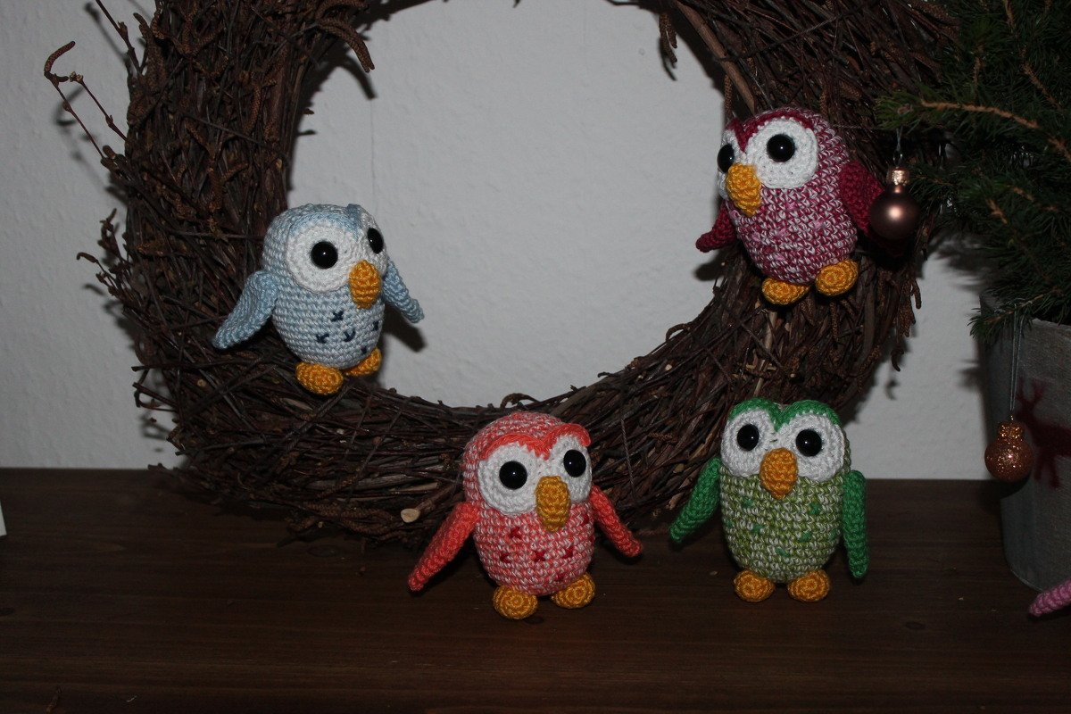 Vier gehäkelte Amigurumi-Eulen (blau, weinrot, orange, grün) auf einem Kranz aus Zweigen.