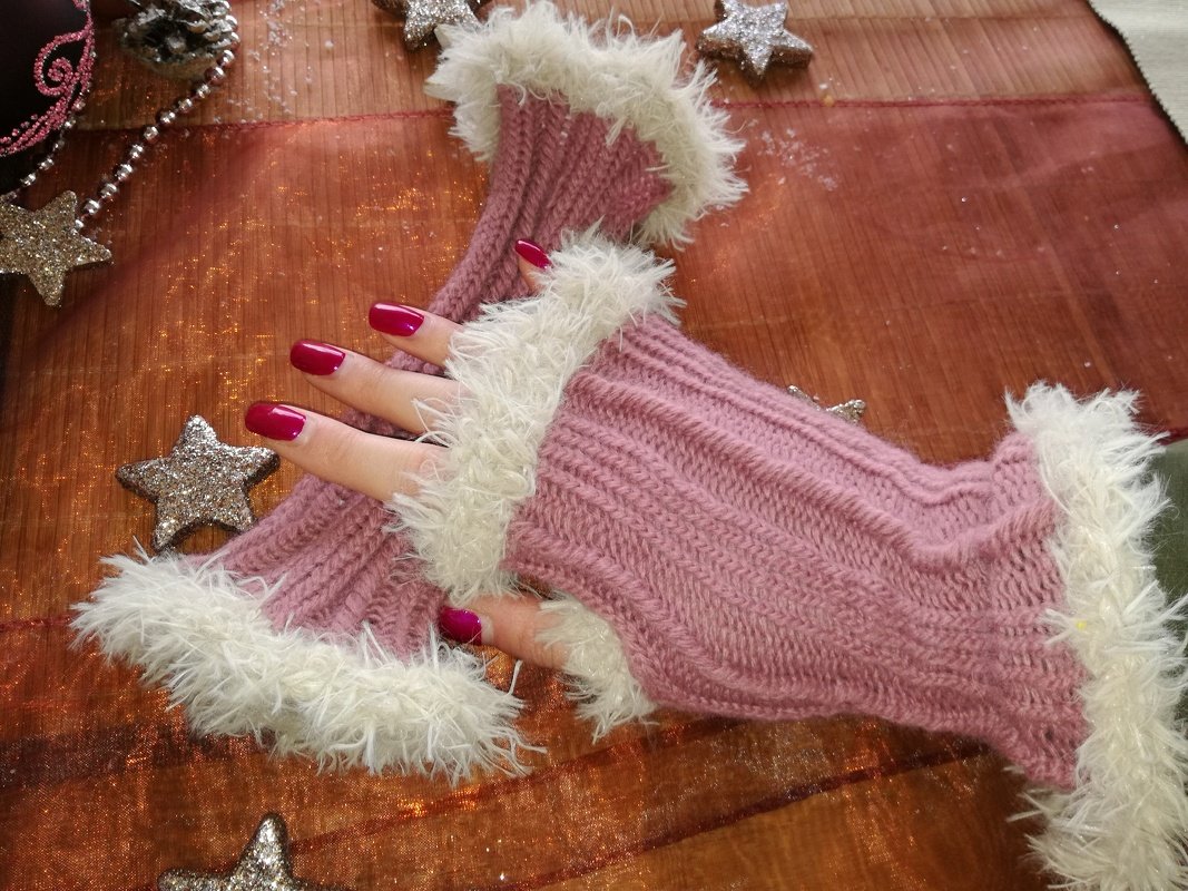 Rosa gestrickte fingerlose Armstulpen mit weißem flauschigem Rand, getragen an Händen mit roten Fingernägeln auf festlichem Untergrund mit glitzernden Sternen.