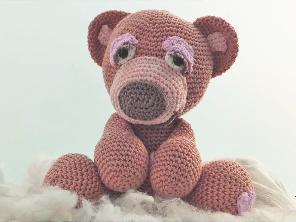 Gehäkelter Amigurumi-Teddybär in Rosa mit lila Akzenten auf weichem Untergrund