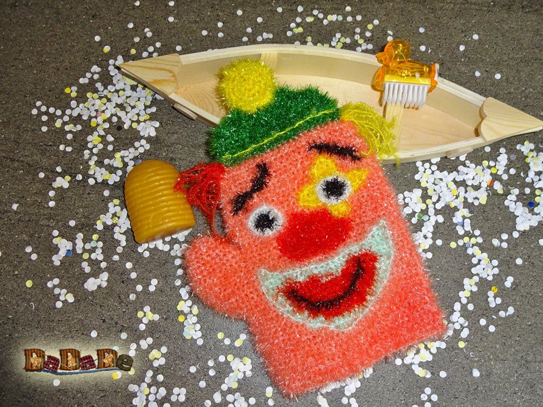 Gehäkelter Clown-Waschhandschuh mit grünem Mützchen, roter Nase, umgeben von Konfetti, Bienenstock-Seife und Holzboot