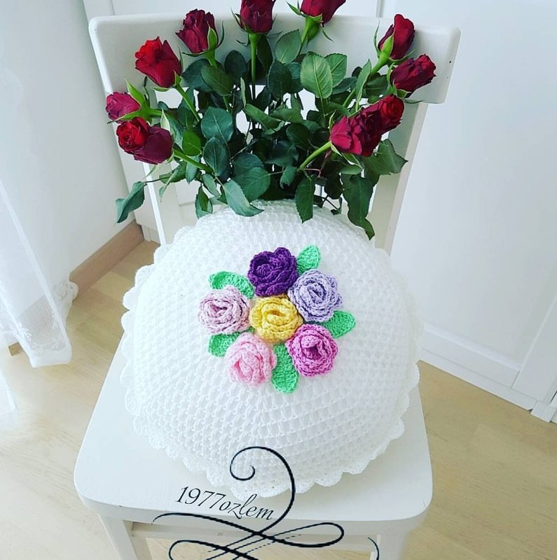 Rundes weißes gehäkeltes Kissen mit bunten Rosen‑Applikationen auf einem Stuhl