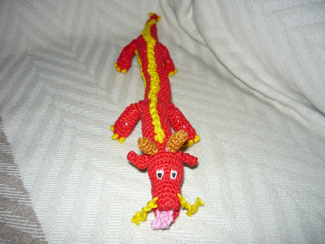 Roter Amigurumi-Drache mit gelbem Rückenkamm und rosa Zunge auf hellem Stoff