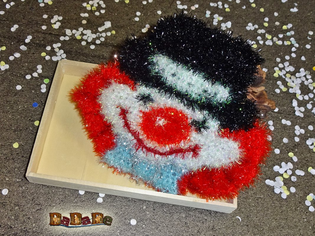 Kompakte, schnell gehäkelte Clown‑Applikation aus flauschigem Effektgarn mit schwarzem Zylinder und roter Nase, auf Holztablett mit Konfetti