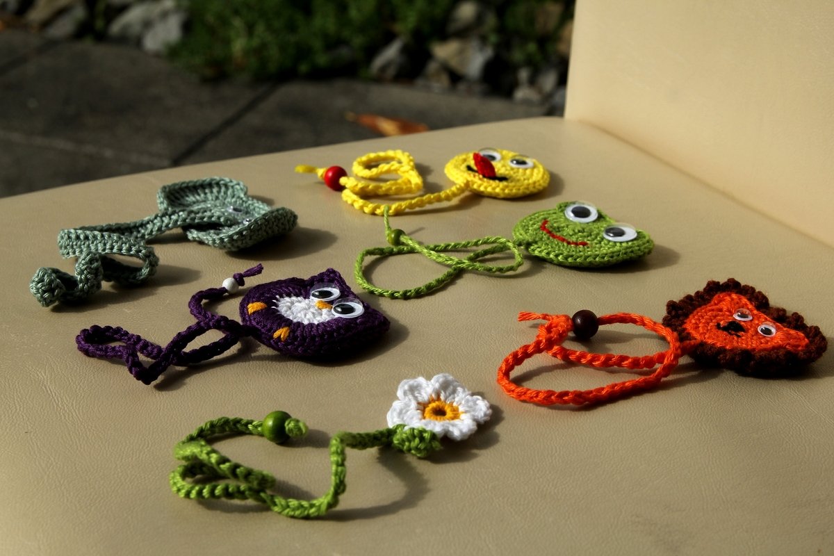 Bunte gehäkelte Lesezeichen in Form von Eule, Frosch, Smiley, Löwe, Blatt und Gänseblümchen auf hellem Untergrund