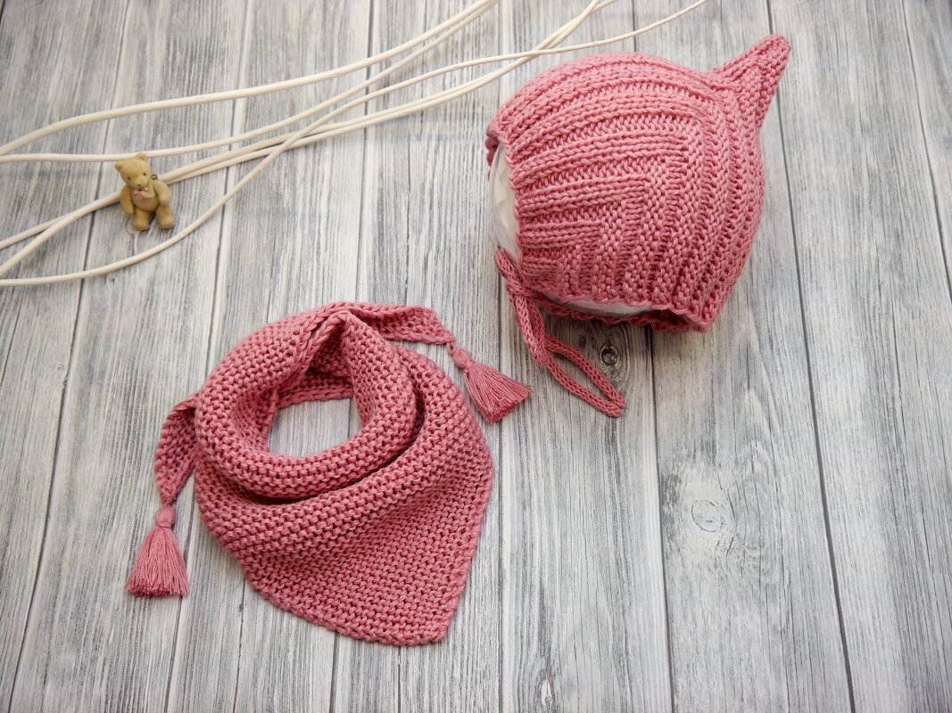 Gestricktes Babyset in Altrosa auf Holzuntergrund: spitze Mütze mit Bindebändern und dreieckiges Halstuch mit Quasten.