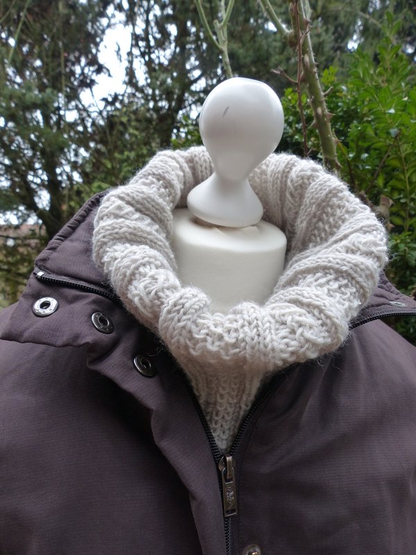 Beigefarbener Strick-Loop auf einer Büste, kombiniert mit dunkler Jacke vor grünem Hintergrund.