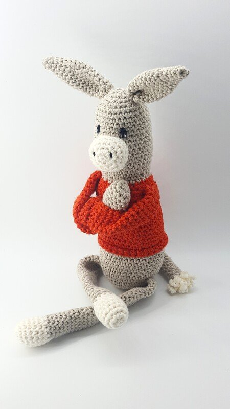 Gehäkelter Amigurumi-Esel mit langen Ohren und orangefarbenem Pullover, sitzend auf weißem Hintergrund.