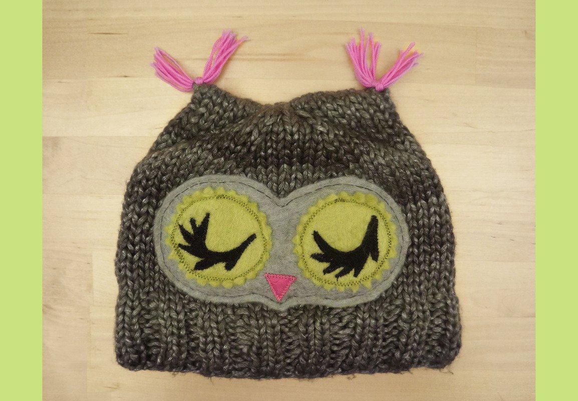 Grau-braune gestrickte Eulenmütze mit gelben Filzaugen, schwarzen Wimpern und pinken Quasten