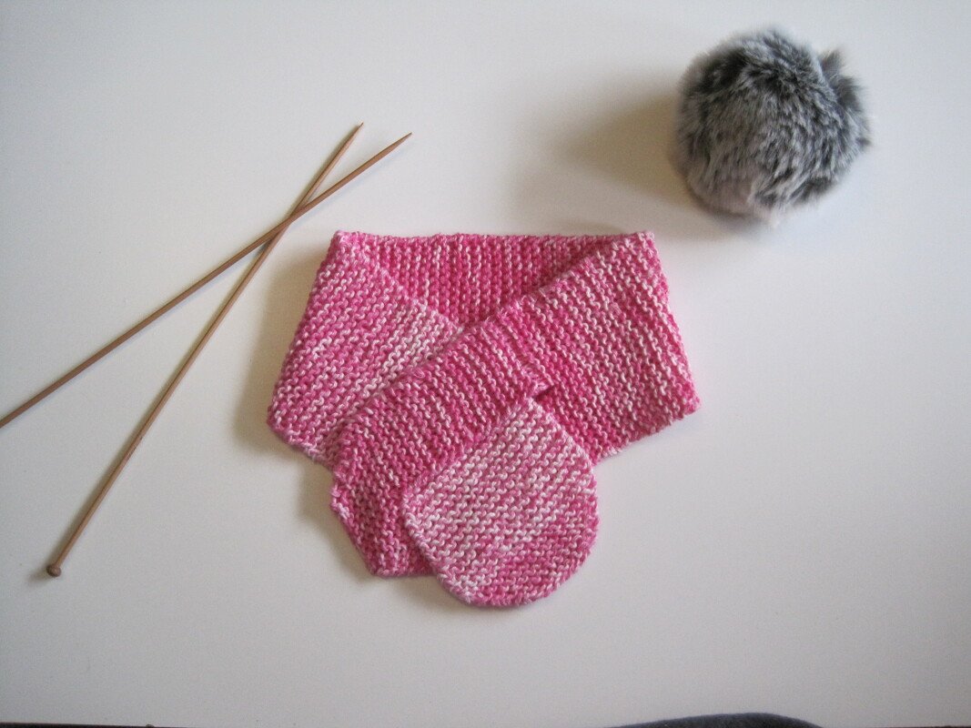 Gestrickter pinker Babyschal neben Holzstricknadeln und grauem Bommel