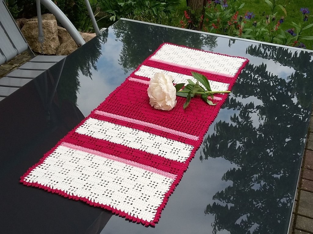 Gehäkelter Tischläufer in Rot und Weiß auf dunklem Glastisch, dekoriert mit einer weißen Pfingstrose.