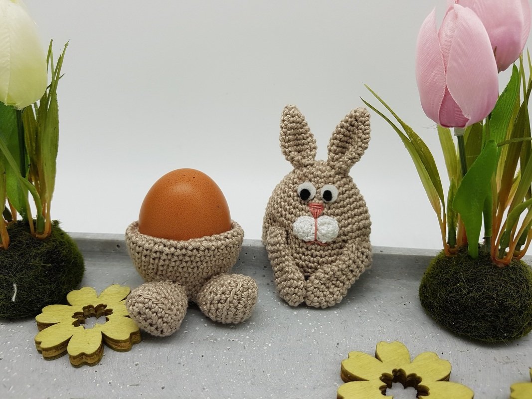 Gehäkelter Eierwärmer in Hasenform neben einem gehäkelten Eierbecher mit Pfoten und braunem Ei
