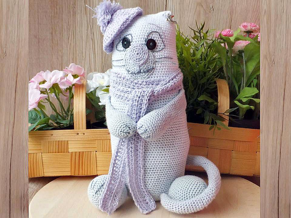 Sitzende gehäkelte Amigurumi-Katze in hellem Flieder mit Bommelmütze und langem Schal vor Holzkörben mit Blumen.