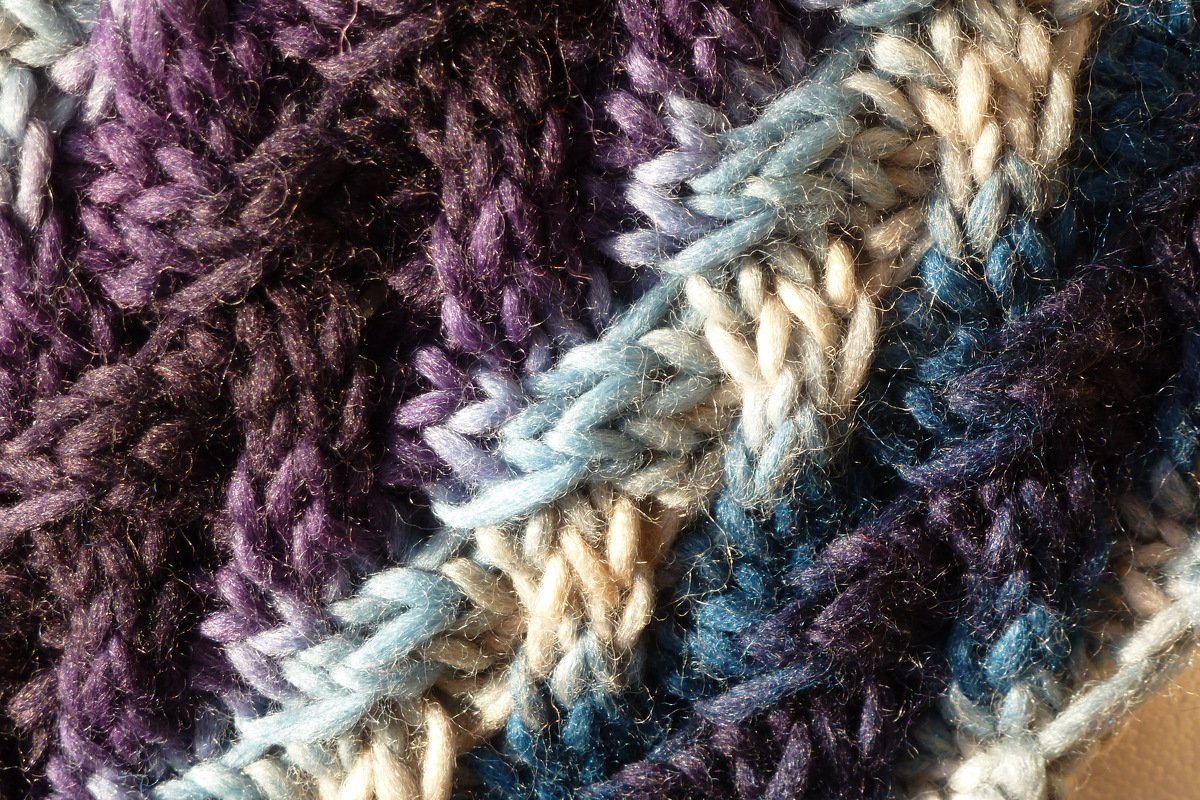Nahaufnahme gestrickter Textur in Violett, Blau und Beige mit zopfartiger, voluminöser Optik.