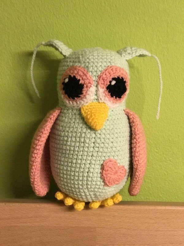 Gehäkelte Amigurumi-Eule in Mintgrün und Rosa mit gelbem Schnabel und gelben Füßen, stehend.