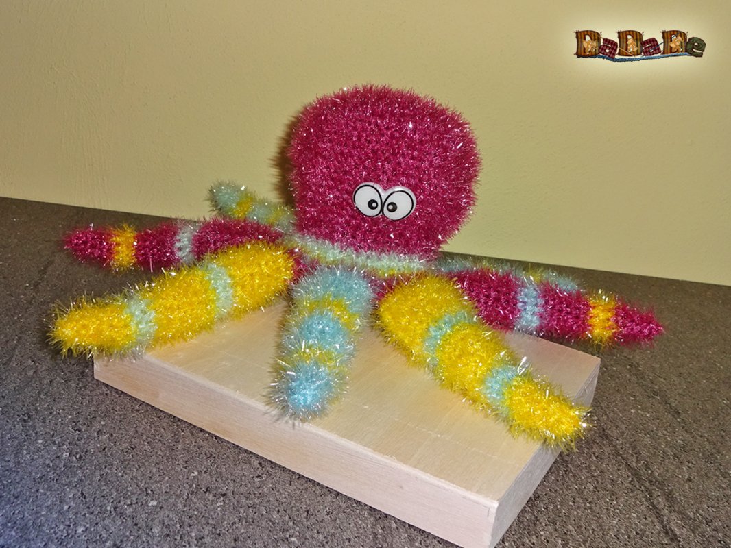 Gehäkelter Amigurumi-Oktopus in Pink, Gelb und Hellblau mit großen Augen auf einer Holzunterlage