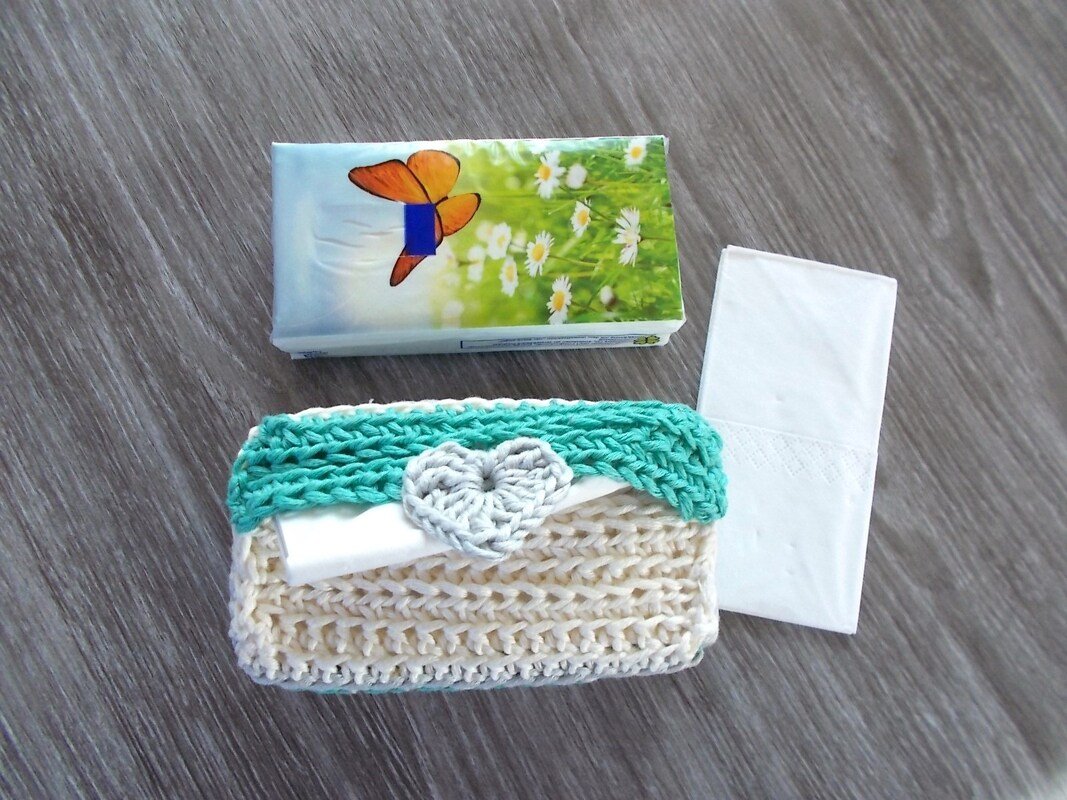 Gehäkelte Taschentücherhülle in Beige mit mintgrünem Rand und grauem Herz auf einer Holzoberfläche, daneben ein Taschentuchpäckchen und ein einzelnes Taschentuch.