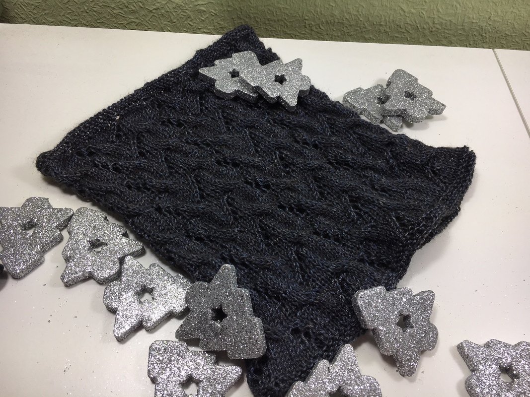 Dunkelgrauer gestrickter Kragenschal mit Zopf- und Lochmuster, dekoriert mit silbernen Sternen auf weißer Unterlage.