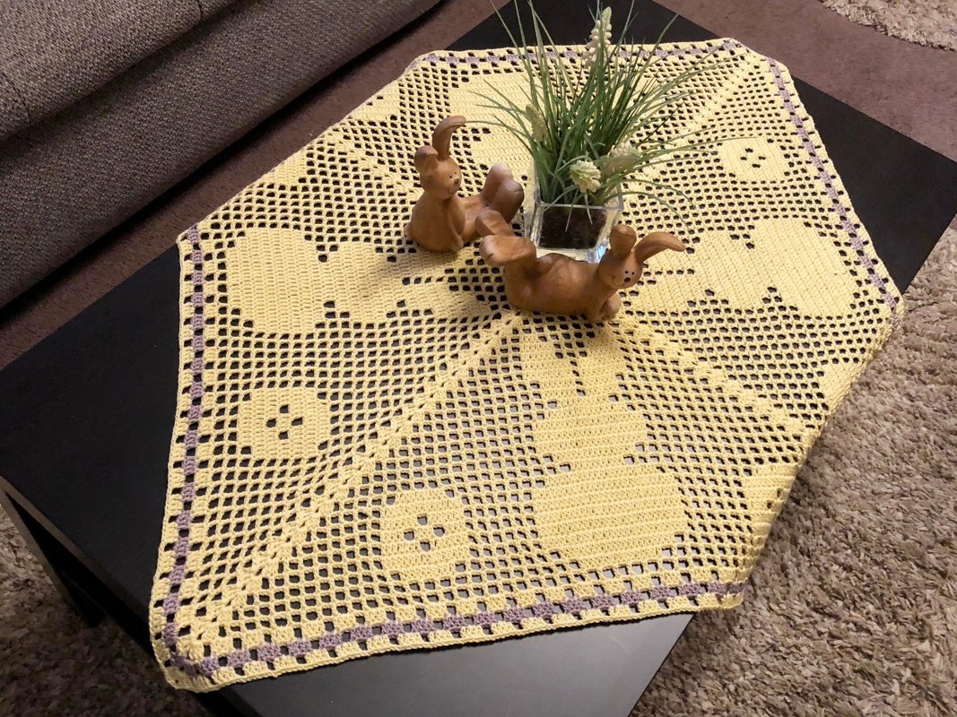 Gelber gehäkelter Tischläufer mit Hasen- und Eimotiven und lila Rand auf dunklem Tisch, dekoriert mit Keramikhasen und Pflanze