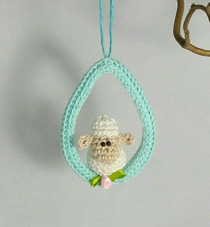 Gehäkeltes Lamm in mintgrünem Eierrahmen mit rosa Blüte und grünen Blättern, an einer Schnur hängend.