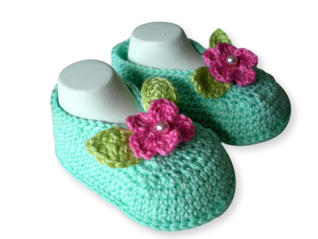 Ein Paar mintgrüner, gehäkelter Babyschuhe mit rosa Blüten und grünen Blättern auf weißen Schuhformen.