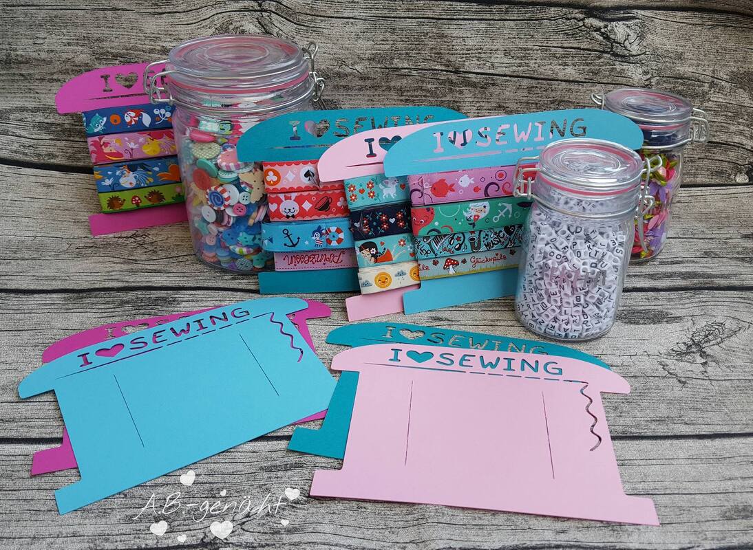 Nahaufnahme von pinken und blauen Bandhaltern in Nähmaschinenform mit Webbändern, umgeben von Gläsern mit Knöpfen und Perlen auf Holzhintergrund.