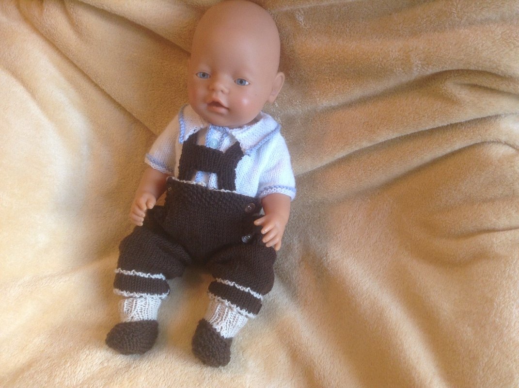 Babypuppe mit brauner gestrickter Latzhose, weißen Söckchen und weißem Kurzarmshirt auf beiger Decke