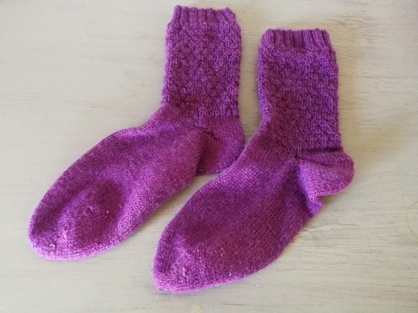 Zwei violette gestrickte Socken nebeneinander auf hellem Untergrund; strukturierter Schaft, glatter Fuß