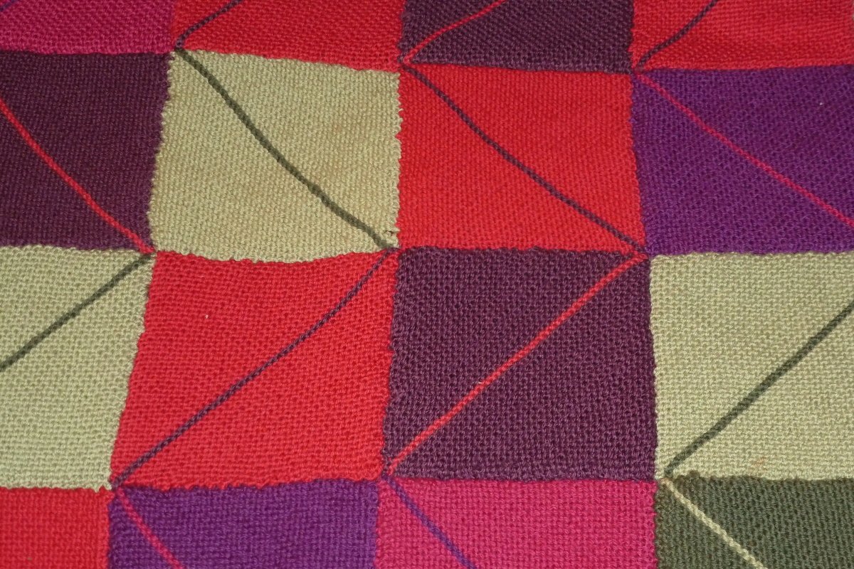 Gestrickte Patchwork‑Decke aus quadratischen Panels in Rot, Magenta, Violett, Beige und Grün mit diagonalen Nahtlinien