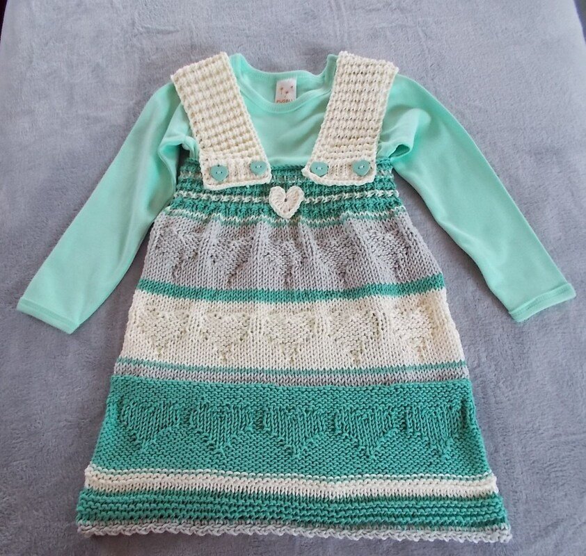 Gestricktes Kinderkleid in Mint, Grau und Creme mit Herzmustern und Knopfträgern, liegend auf hellem Untergrund