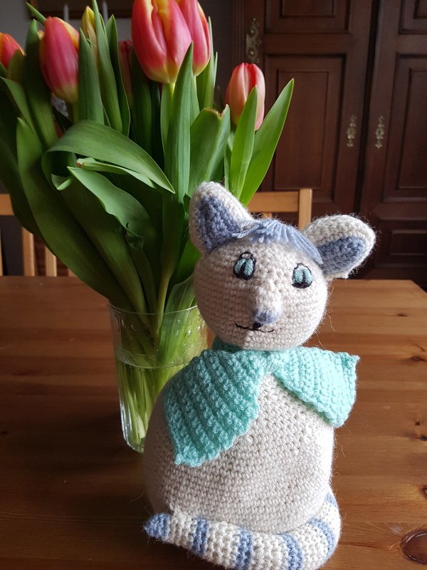 Gehäkelte Amigurumi-Katze in Creme mit hellblauen Akzenten und Schal, sitzend auf einem Holztisch neben einer Vase mit Tulpen.