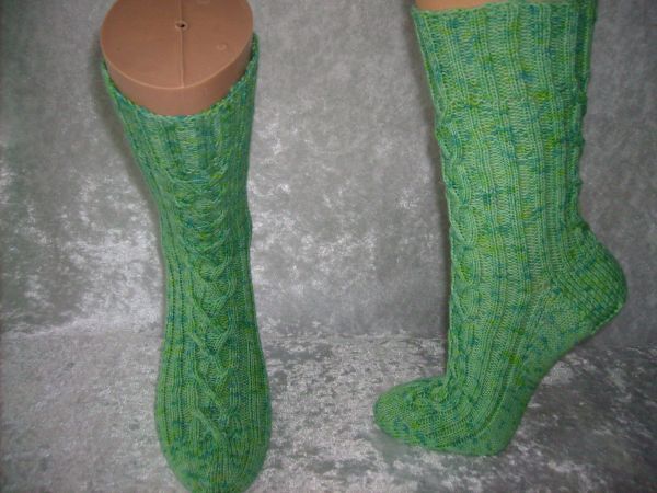 Gestrickte grüne Socken mit Zopfmuster auf Mannequin-Beinen vor neutralem Hintergrund.