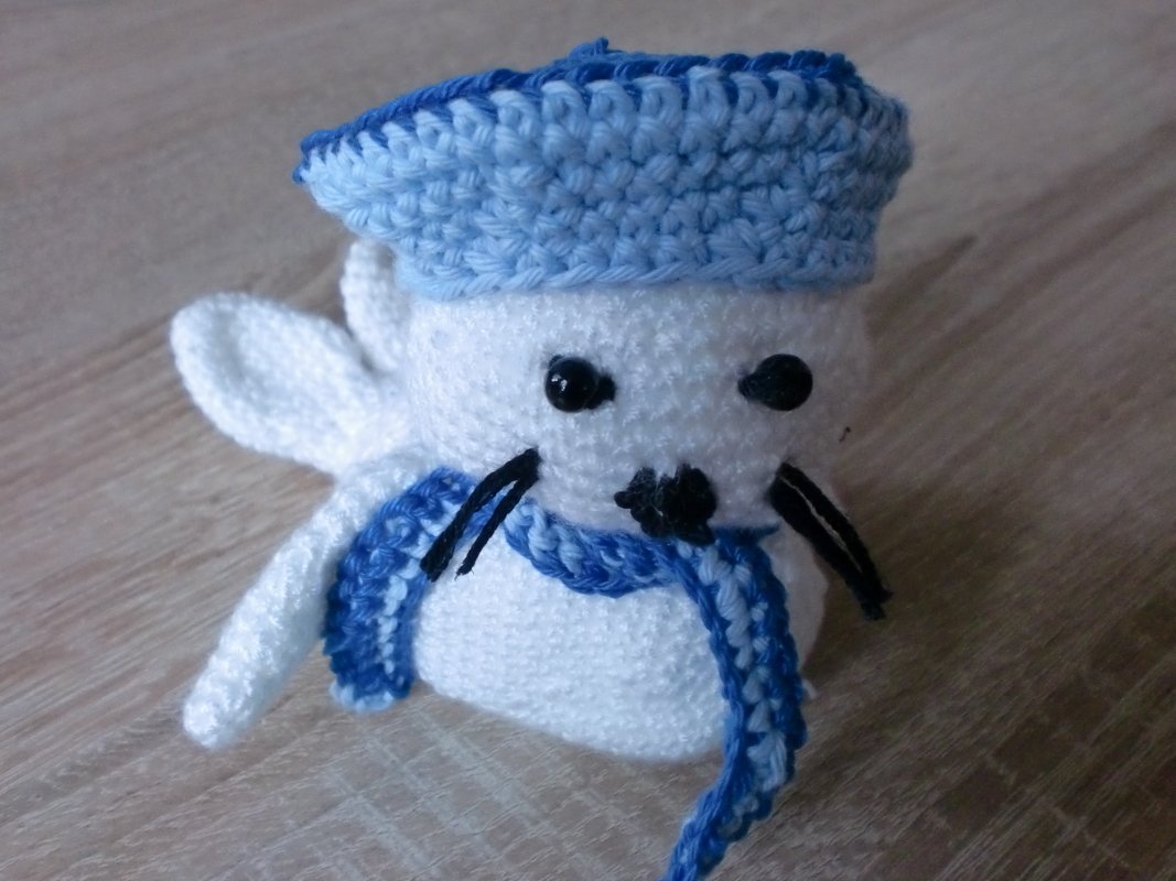 Weißer gehäkelter Seehund Amigurumi mit blauer Matrosenmütze und Schal auf Holzuntergrund