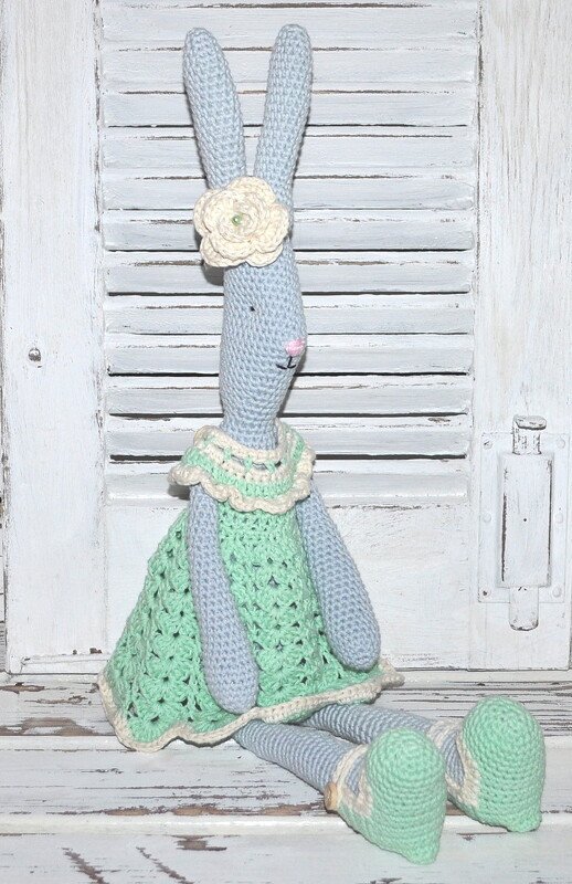 Sitzender gehäkelter Hase in Hellblau mit mintgrünem Kleid und cremefarbener Blüte vor weißem Holzlattenhintergrund.