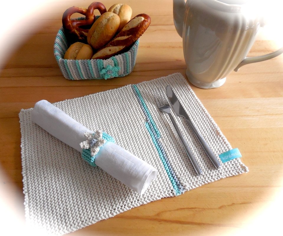 Gestricktes cremeweißes Tischset mit türkisen Akzenten, Serviettenring mit Blüte und gestreifter Brotkorb mit Backwaren.