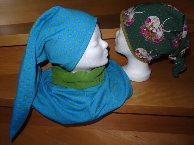 Zwei genähte Zipfelmützen auf Modellköpfen: eine blaue mit Sternen und Loop-Schal, die andere dunkelgrün mit Hirsch- und Rosenmuster.