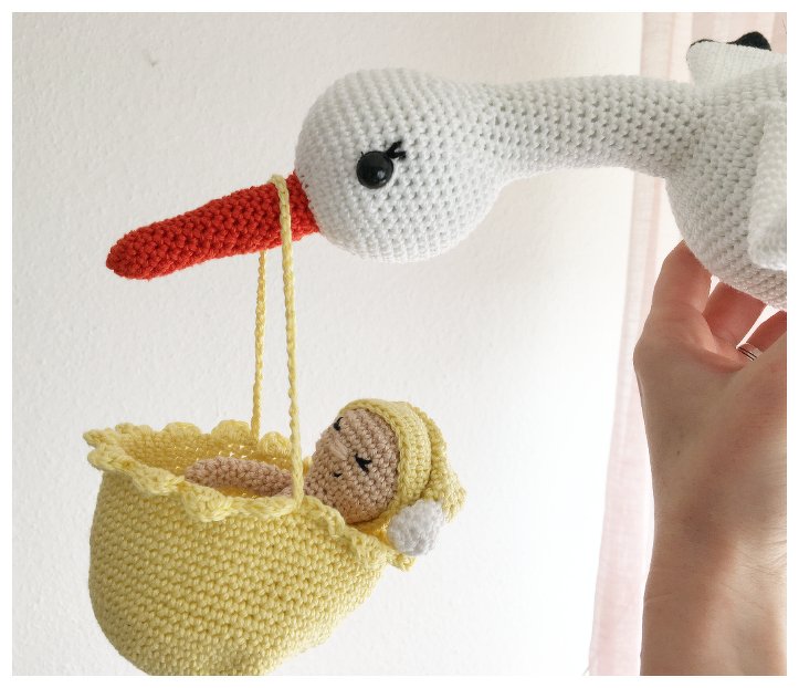 Gehäkelter weißer Storch mit orangefarbenem Schnabel hält gelbes Körbchen mit gehäkeltem Baby