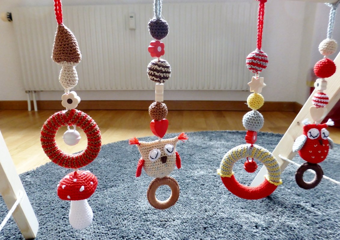 Detailansicht von vier handgehäkelten Anhängern am Baby-Spieltrapez, darunter Pilze, Eulen und Greifringe. Mit Holzperlen und Glöckchen in Naturtönen.