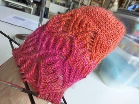 Nahaufnahme einer handgestrickten Socke in Pink und Orange mit Lochmuster auf Stricknadeln.