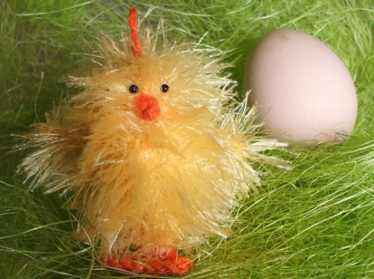 Gelbes flauschiges Strickküken mit orangem Schnabel und Füßen neben einem hellen Ei auf grünem Dekogras.