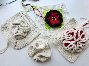 Nahaufnahme mehrerer gehäkelter Granny Squares mit roter Mohnblüte, 3D-Rüschen und losem Garn
