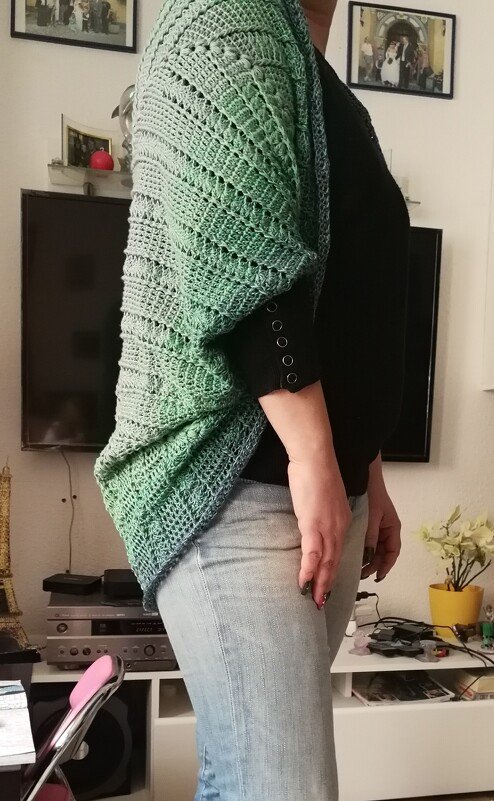 Seitenansicht: gehäkelter Seelenwärmer in Grün‑Blau über schwarzem Top und Jeans, mit durchbrochenem Rauten‑ und Wellenmuster.