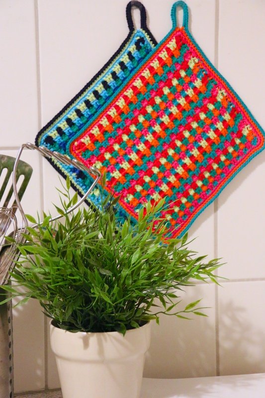 Zwei farbenfrohe gehäkelte Topflappen im Granny‑Square‑Muster mit Aufhängeschlaufen neben einer grünen Topfpflanze.