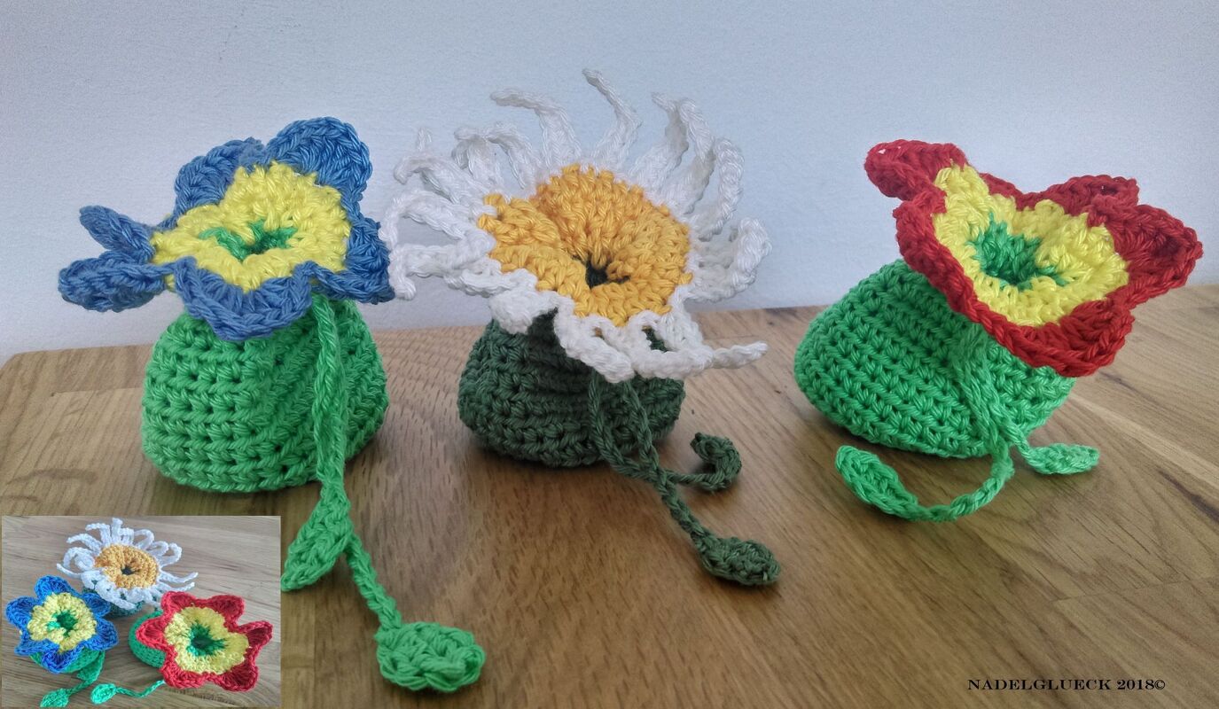 Drei gehäkelte Blüten-Täschchen: grüne Beutel mit blau‑gelber, weiß‑gelber und rot‑gelber Blüte